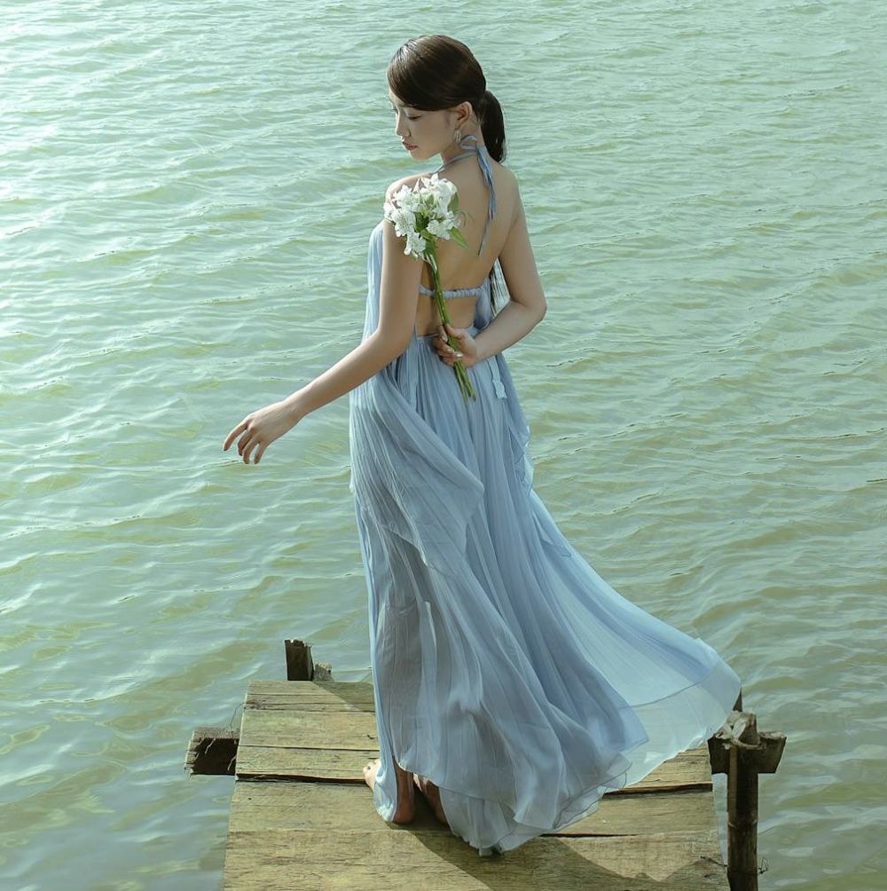 young asian woman in a flowing dress, holding flowers on her back, waves background